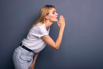 Caucasian woman in neutral casual outfit standing on a neutral grey background. Portrait with emotions: happiness, amazement, joy and satisfaction while whispering secrets