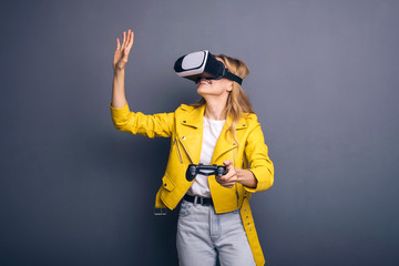 Fototapeta na wymiar Caucasian woman in neutral casual outfit standing on a neutral grey background. She playing game using joystick with VR goggles and smiling happily