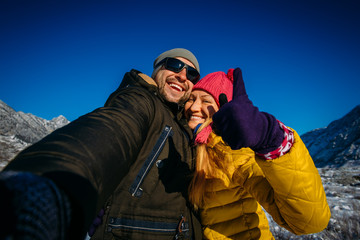 Guy and girl embrace and smile on the background of snow-capped mountains and blue sky. Happy young couple taking selfie during winter vacation in mountains.