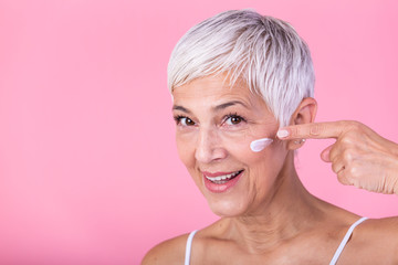 Beautiful mature woman holding jar of skin cream for face and body isolated on pink background....