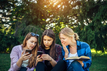 Three cute girls relax and socialize on the lawn in the summer park. Young women on the green grass among the trees, looking at smartphone, reading a book. Students in between classes outdoor.