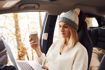 Sits with laptop and cup of drink. Girl have autumn trip by car. Modern brand new automobile in the forest