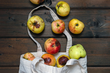 Zero waste concept. Red and yellow ugly apples in white textile bag on wooden background. apples scattered on the table. view from above.