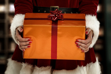 Closeup of orange gift with red ribbon and bow.Woman hands and red christmas dress.Home interior and blurred wall background.Copy space.Free space for your decoration.Xmas time and cold december night