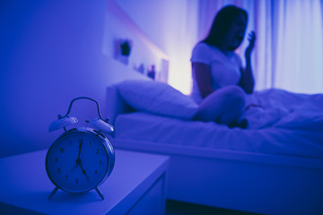 Close-up view of alarm call bell clock nice attractive tired hardworking girl sitting in bed yawning life regime bedtime in dark blue illuminated room house hotel flat indoors