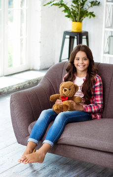 That’s my Teddy bear. Full-length photo of beautiful little girl with long wavy chestnut hair, looking into camera, smiling broadly and holding a teddy bear while sitting on the couch.