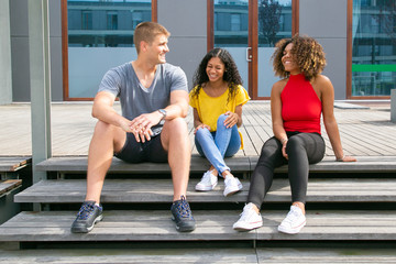 Smiling friends talking outdoors. Happy young multiethnic friends sitting on wooden surface and talking on street. Communication concept