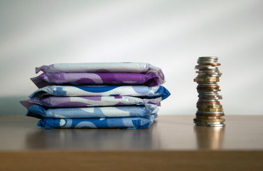 A stack of sanitary towels next to a taller stack of coins. Illustrating period poverty and...