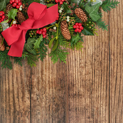 Festive Christmas, winter & New Year  background border with a red bow,  holly, snow covered fir, mistletoe, pine cones, ivy & cedar leaves on rustic wood with copy space.