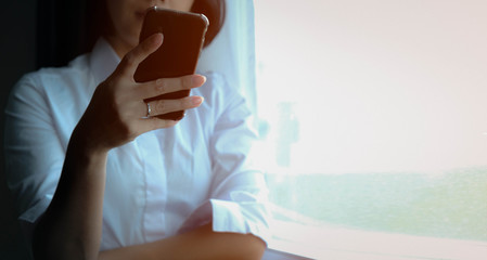 Asian woman, short black hair wear white shirt standing beside a window with some light passing through. In hand holding a mobile phone and a little smile. Maybe shopping online or chat to her lovers.