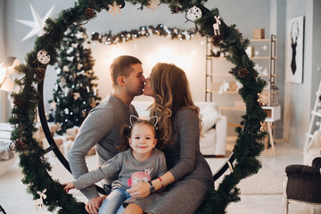 Full length stock photo of three family members daughter, mother and father snuggling on decorated Christmas swing in circled shape with fir branches.