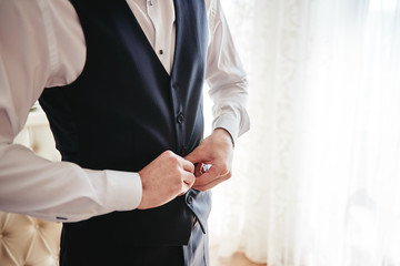 Man in white shirt buttoning waistcoat in room indoors, close up. Businessman dressing up. Fashion,...