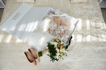 Luxury wedding dress in white box, beige women's shoes and bridal bouquet on bed, copy space. Bridal morning preparations. Wedding concept