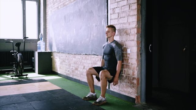 Male fitness instructor does wall sit exercise at gym. Wall sits to make hip and thigh stronger. Effective exercises for running. Short flexibility-focus session at workout place.