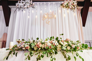 Wedding presidium in restaurant, copy space. Banquet table for newlyweds with flowers, greenery, candles and garland ligths. Lush floral arrangement. Luxury wedding decorations