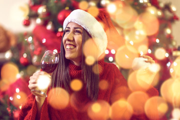 young woman in Santa hat by Christmas tree at home toasting with red wine.