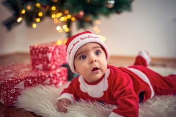 Cute baby in Christmas costume and gift box on floor at home