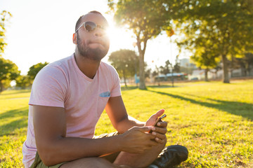 Naklejka na ściany i meble Peaceful handsome guy with cellphone enjoying leisure time outdoors. Latin man sitting on grass at sunset in park, holding smartphone, looking away. Leisure concept