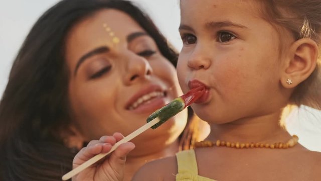 Close up view of smiling indian woman enjoys the moment with her pleased child girl which eating lollipop in park outdoors - Powered by Adobe