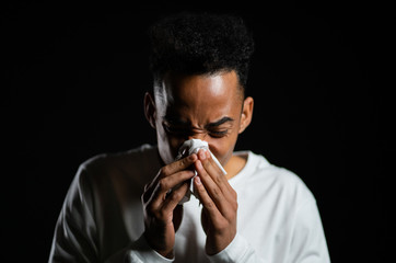 Young african american man sneezes into tissue. Isolated man is sick, has a cold or has allergic reaction. Health, medicine, illness, treatment concept