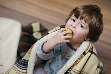 Portrait of a small curly-haired boy. A child wrapped in a rug eats an apple. A two-year-old boy hides under a blanket.