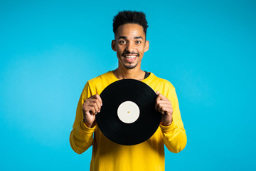 Young handsome african american man in yellow wear holding vinyl records at blue background. Retro...