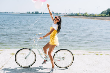 Fototapeta na wymiar side view of happy brunette girl riding bike with balloons near river in summer