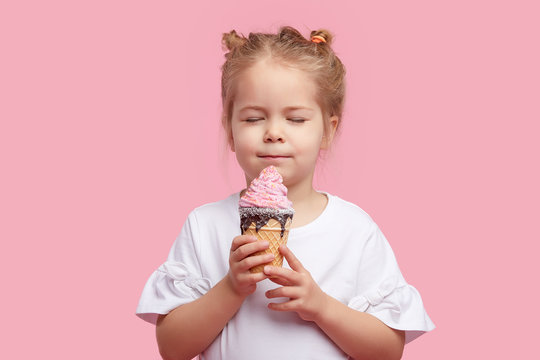 Cute Child Girl With Pleasure Eats Tasty Ice-cream On A Pink Studio Background. Licks With Closed Eyes. The Concept Of Baby Food And A Happy Childhood