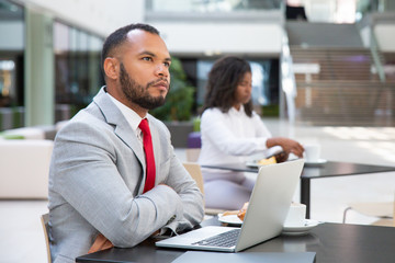 Pensive businessman with laptop thinking over startup strategy over cup of coffee. Young African American woman drinking coffee in background. Working in cafe concept