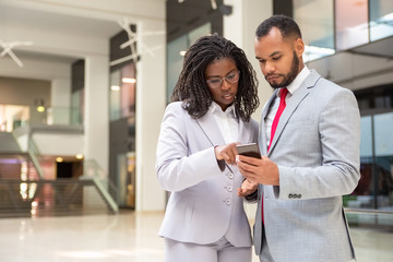 Focused diverse colleagues using mobile phone together. Business man and woman wearing formal suits, holding smartphone and pointing at screen. Watching content concept