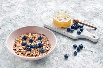 Muesli or granola with blueberries and jar of fresh honey and stick on wooden board, grey textured background. Healthy, vegan, raw food  concept. Beautiful oatmeal breakfast, selective focus,