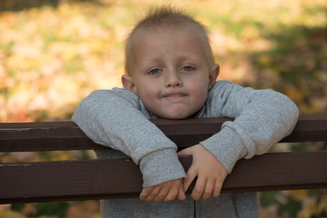 little blond boy leaning on the back of a bench in the park and smiling