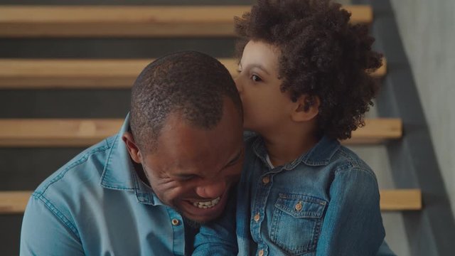 Cheerful smiling adorable mixed race preschool son whispering gossips, sharing secrets with handsome laughing black father on stairs at home. Trust-based warm relations between daddy and child.