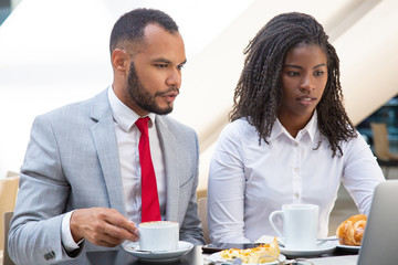 Focused diverse colleagues working on laptop together during breakfast. Business man and woman drinking coffee in cafe, using computer, looking at screen. Wireless technology concept