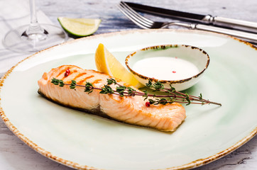 Baked Salmon Steak with herbs, sauce and lemon slice on plate. Delicious fried fish served coarse salt and cherry tomato branch for dinner.