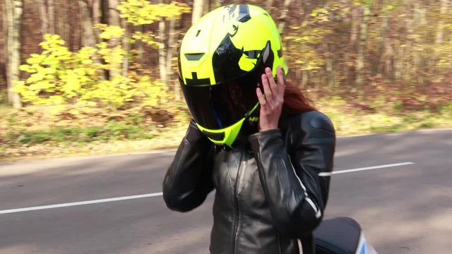 The red-haired girl in a leather jacket takes off a neon motorcycle helmet. Riding. Racer. Sport motorcycle. Autumn forest. Travel.