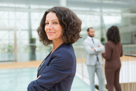 Friendly successful business woman posing with arms crossed, looking at camera, smiling. Her male and female colleagues standing and talking in background. Female business leader concept - Powered by Adobe
