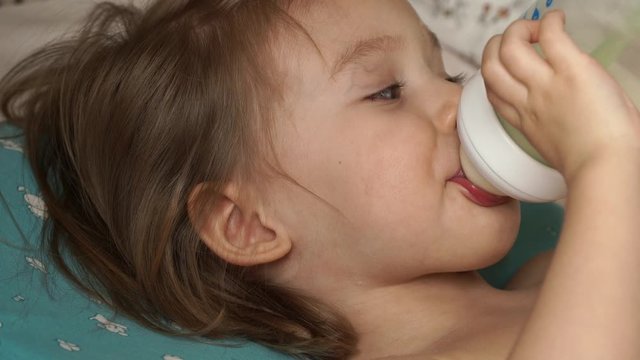 Portrait of little cute baby girl lies after sleep in morning and holds bottle of milk, sucks with great appetite, looks at camera and smile. Infant formula milk concept. Сlose up