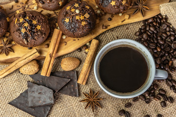 Chocolate holiday cupcakes, muffins with cake sprinkles on a wooden board, a cup of coffee, chocolate, cinnamon sticks, star anise, cocoa, coffee beans, nuts,  burlap sack