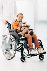 partial view of pediatrist in white coat and kid with teddy bear on wheelchair