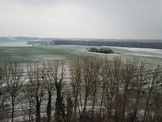vue aérienne d'un champ d'herbe verte à moité sous la neige avec forêt d'hiver en fond  et...
