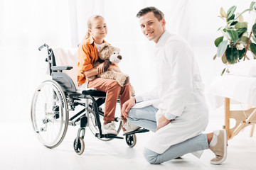 pediatrist in white coat and kid with teddy bear on wheelchair