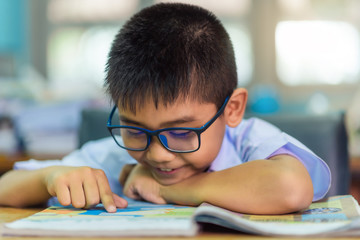 Asian elementary school boy in a white school uniform and wearing glasses, is reading a book in the classroom.