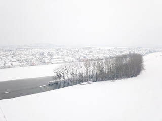 lac gelé et arbres sur champ de neige avec ville de campagne au loin dans une ambiance hivernale de noël par temps neigeux presque tempête de neige vue aérienne