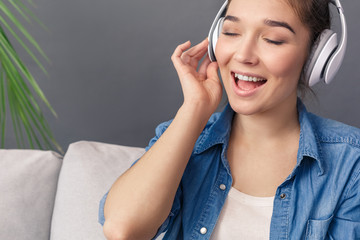 Hobby Concept. Young woman in headphones sitting on sofa studio isolated on grey listening music singing along smiling delightful close-up