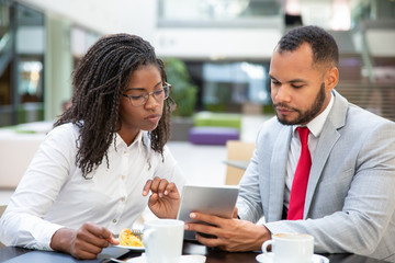 Focused mix raced colleagues watching content on tablet together during lunch. Business man and woman drinking coffee in cafe, using digital device, looking at screen. Lunch with colleague concept