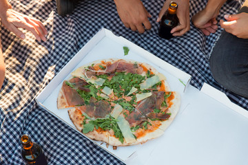 Group of friends eating pizza outdoors. Top view of pizza in box on plaid, beer bottles and human hands. Picnic concept