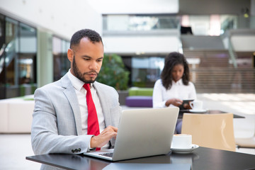 Serious businessman drinking coffee and working on laptop in office lobby. Young African American woman using tablet in background. Public internet connection concept