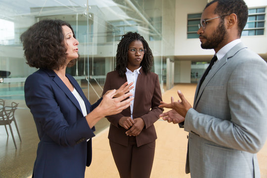 Business Colleagues Discussing Project Near Office Building. Serious Business Man And Women Standing At Outdoor Glass Wall, Talking To Each Other, Gesturing. Corporate Discussion Concept