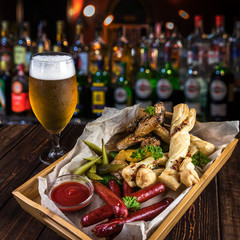 Oktoberfest food snacks concept with blurred background of bar counter. Wooden table with a glass of cold beer, water droplets and a big box with bread, grilled sausages, pickles, chicken, potatoes